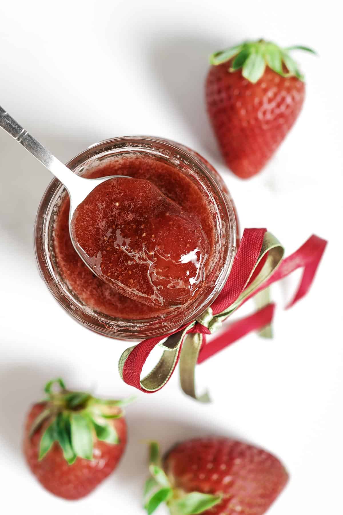 A scoop of strawberry jam in a spoon over a jar of jam.