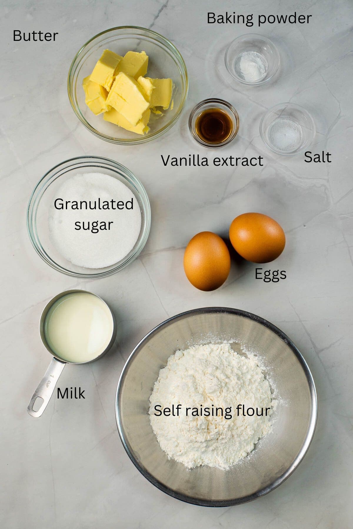 Butter, vanilla extract, salt, sugar, eggs, milk, self raising flour and baking powder against a marble background.