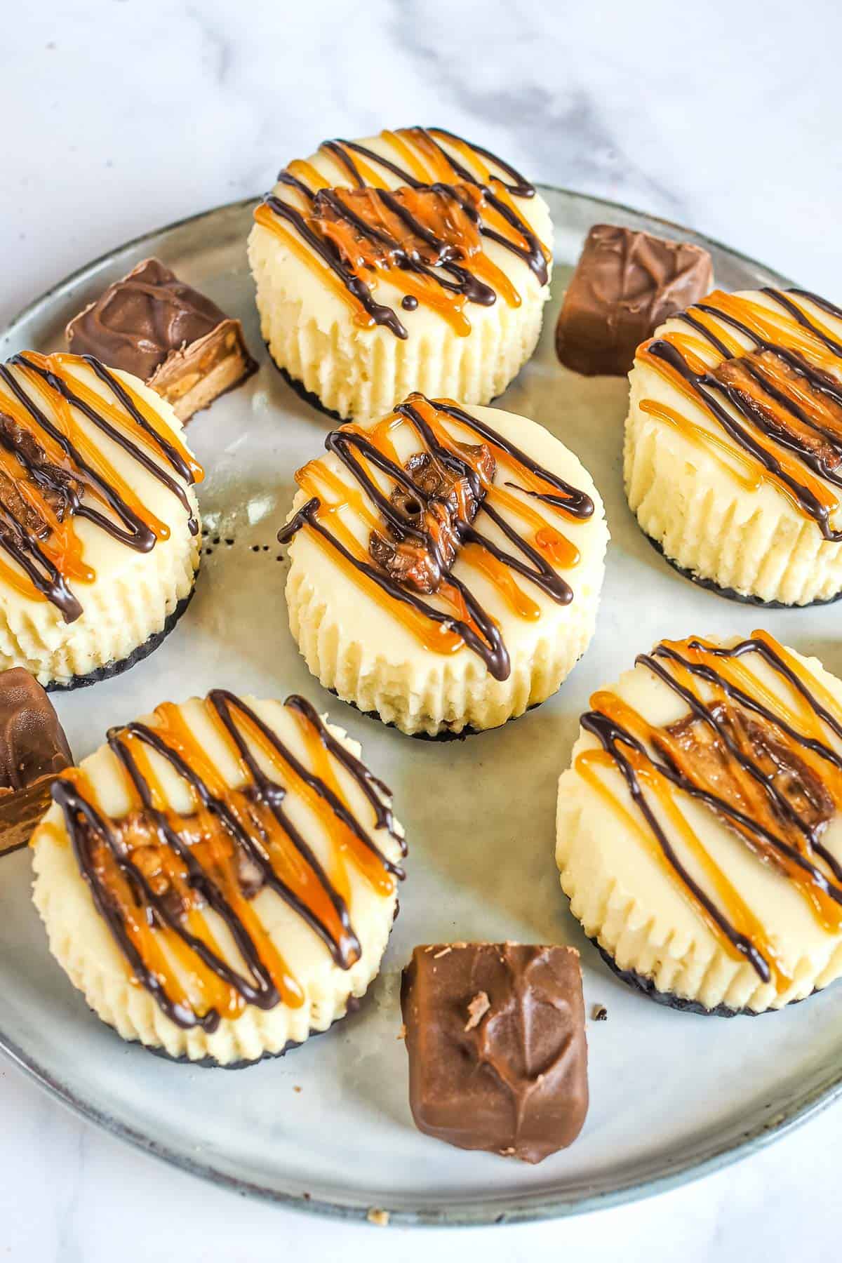 6 mini cheesecakes decorated with Snickers chocolate and chocolate and caramel drizzles on a grey plate