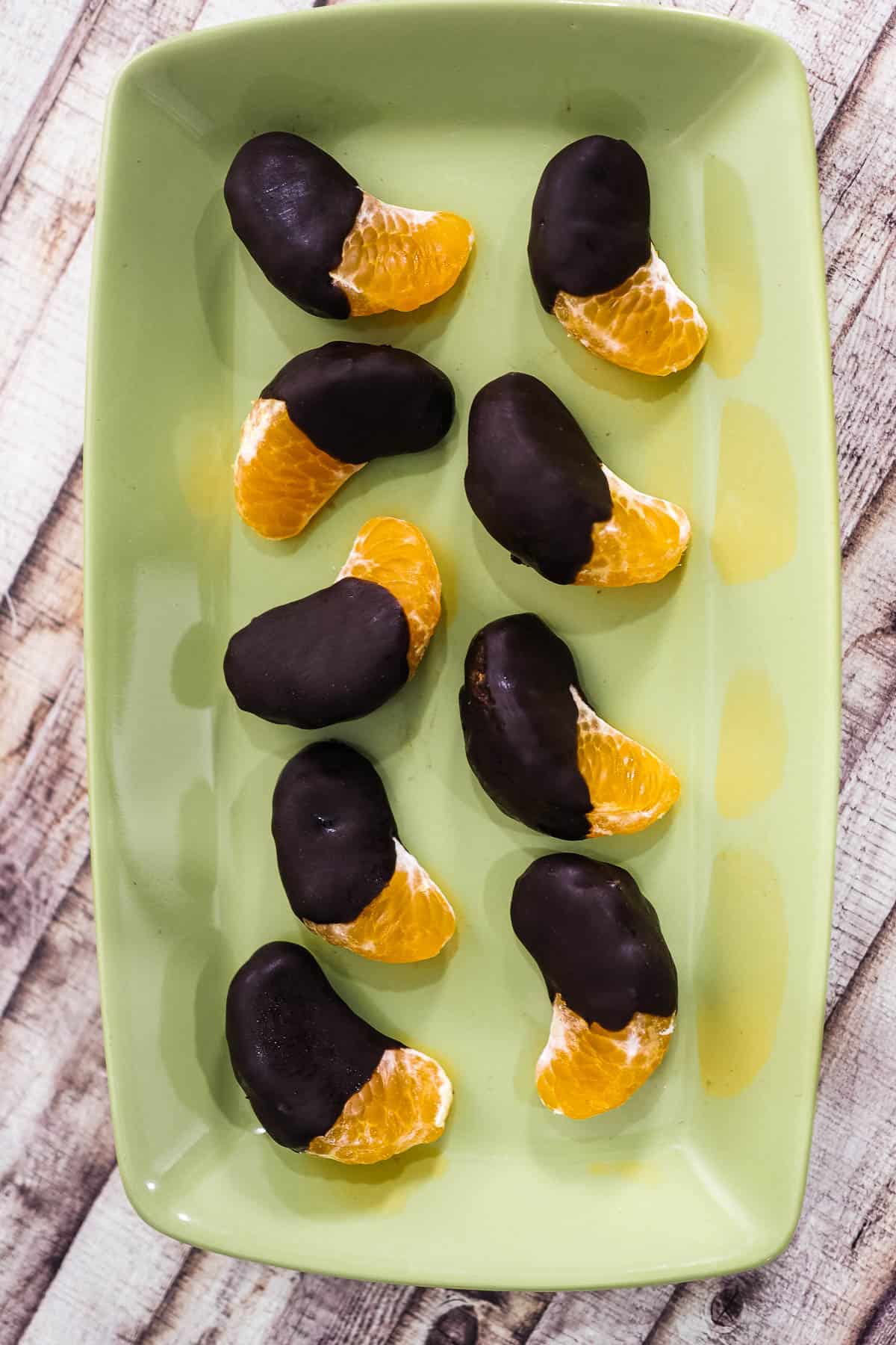 Chocolate dipped orange segments on a green rectangle plate against a white wood background