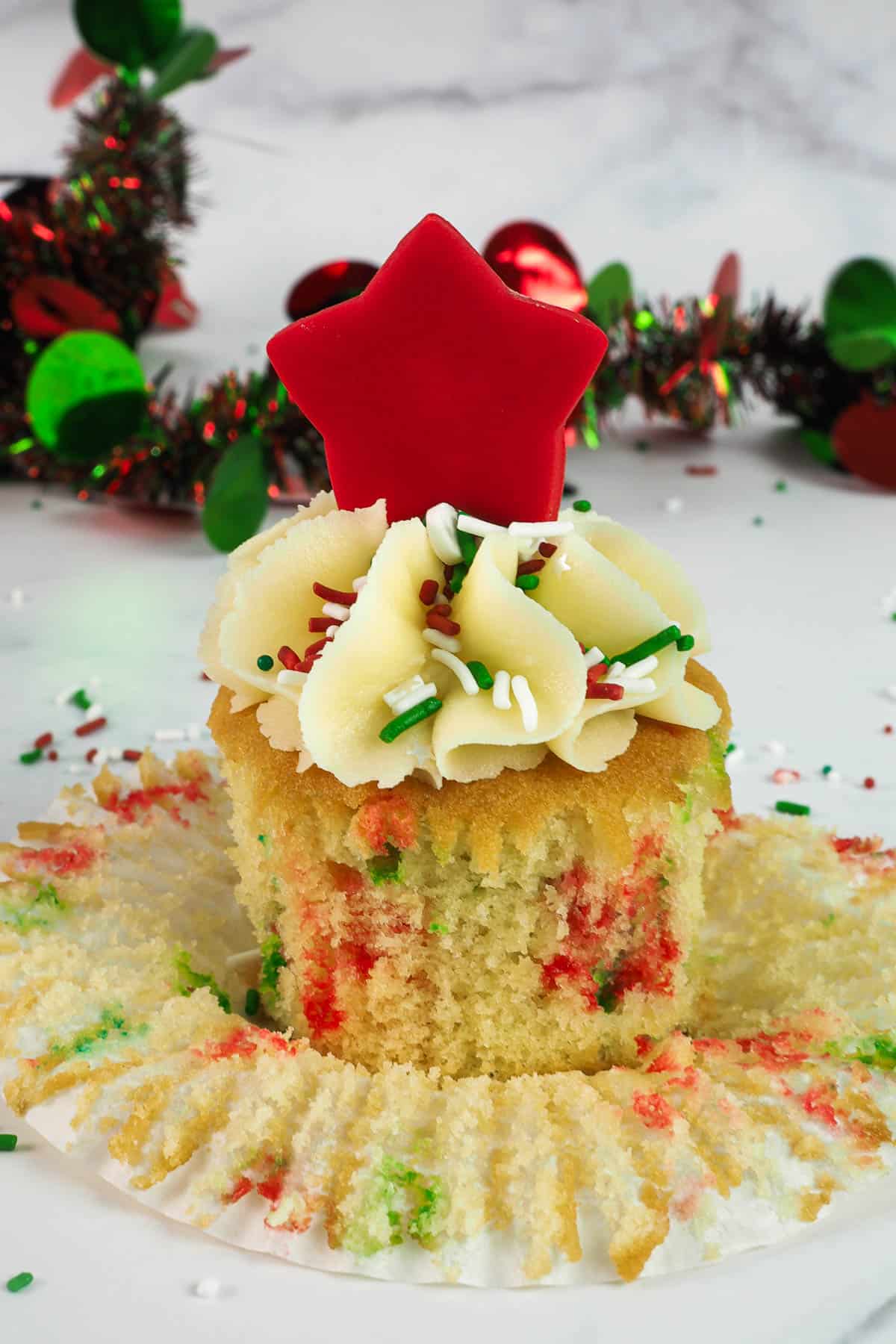 A sprinkles cupcake with its casing peeled off. Cupcake is decorated with simple buttercream swirl, a red star topper and red and green sprinkles.