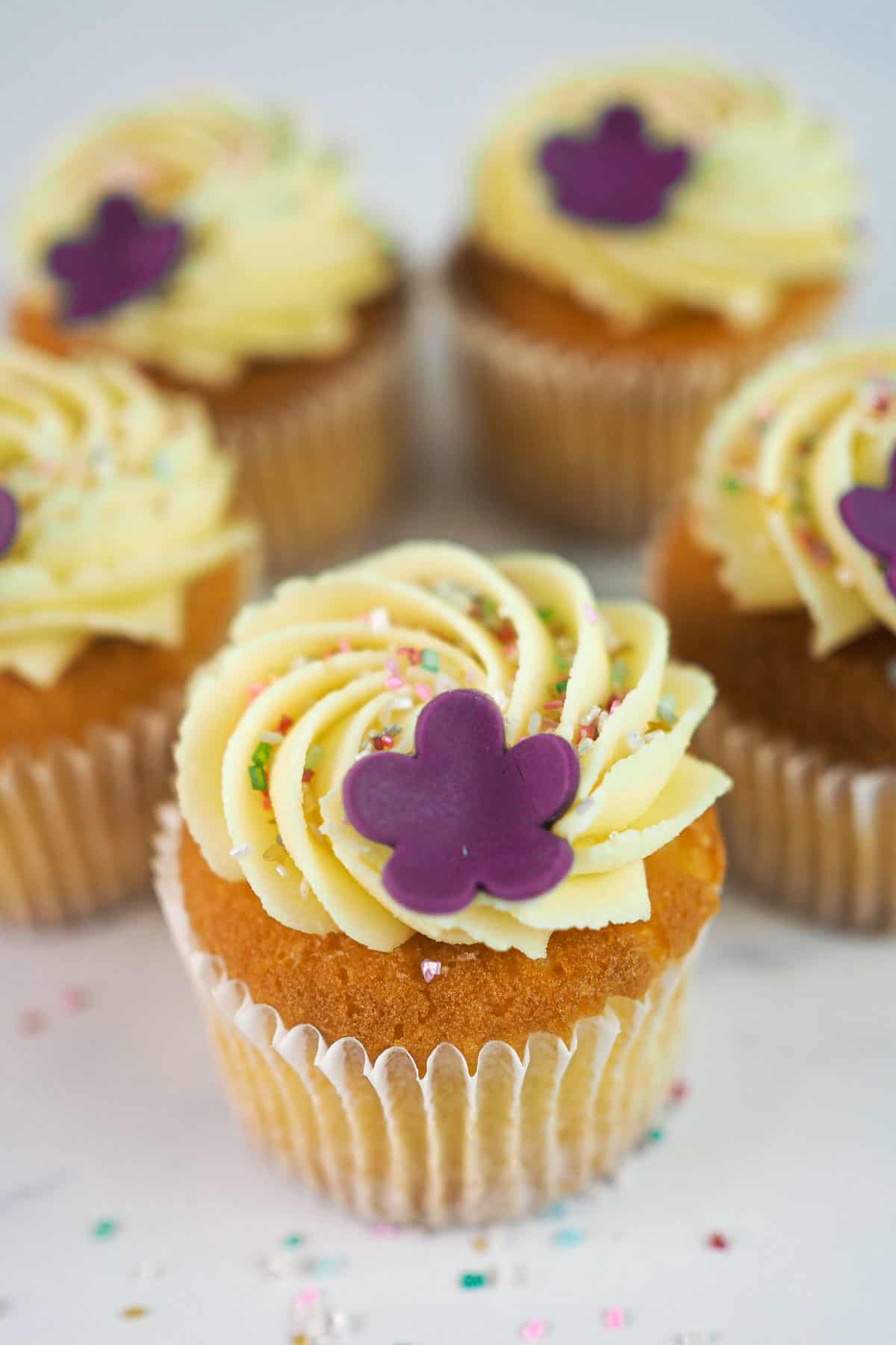 A set of cupcakes in white cupcake casings decorated with buttercream, sugar sprinkles and a pretty 5 petal fondant flower.