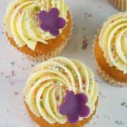 A set of cupcakes in white cupcake casings decorated with buttercream, sugar sprinkles and a pretty 5 petal fondant flower.