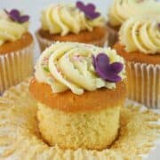 A cupcake with its casing peeled open. Cupcake is decorated with a simple buttercream swirl, sugar sprinkles and a purple 5 petal fondant flower.
