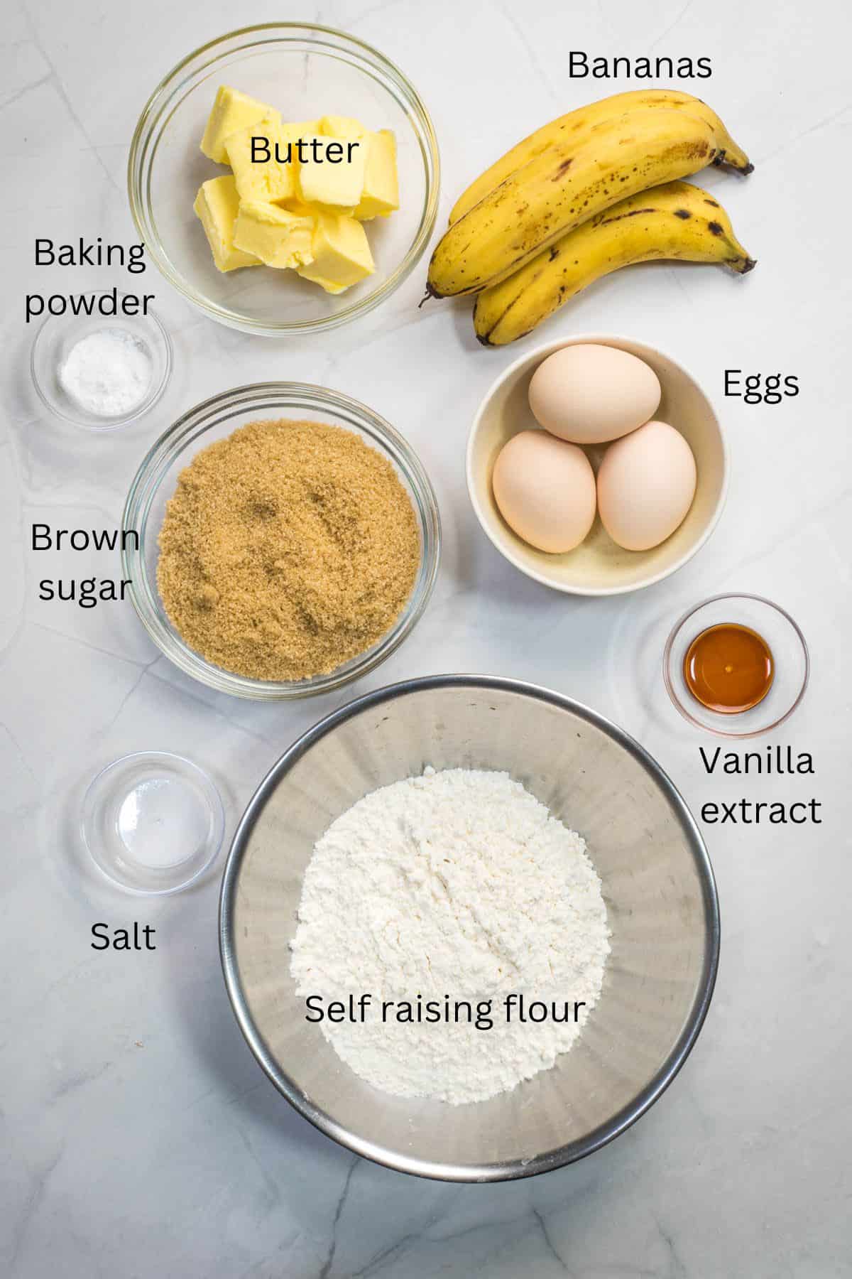 Self raising flour, brown sugar, butter, eggs, vanilla extract, baking powder, salt and bananas against a marble backdrop.