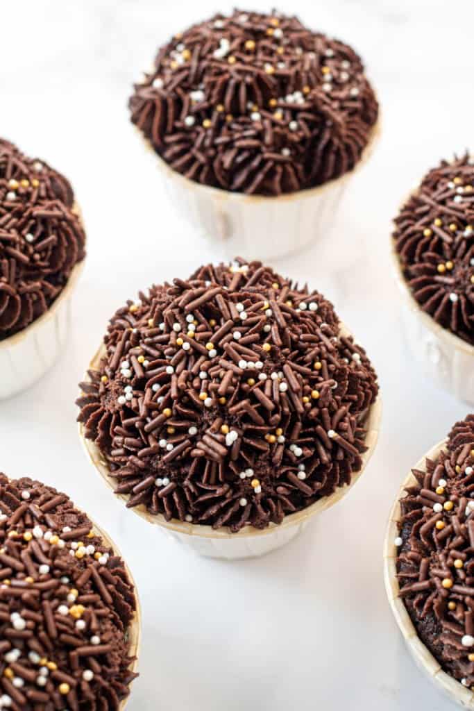 Chocolate cupcake with chocolate buttercream in white cupcake casings. Cupcakes are topped with gold and chocolate sprinkles.