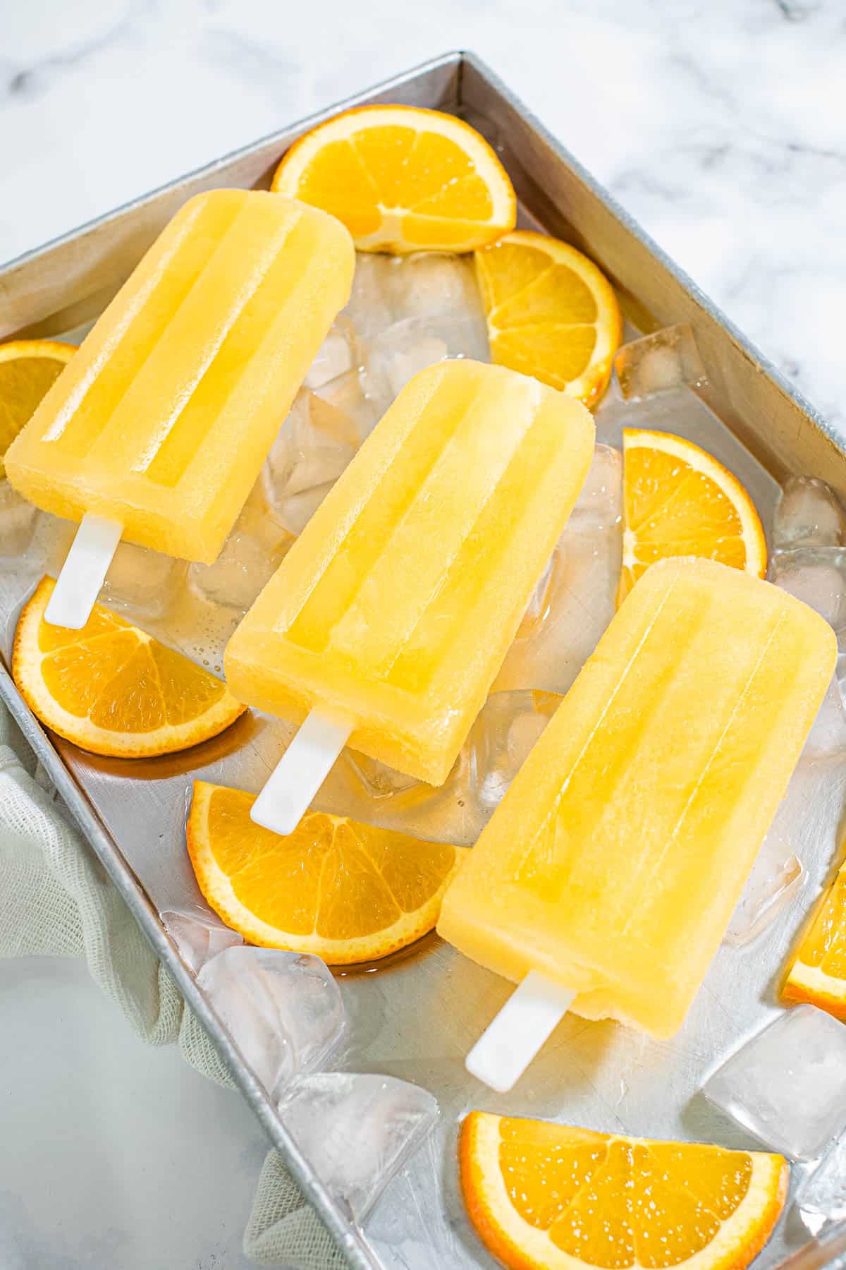 3 orange ice pops on a tray filled with ice cubes and orange slices