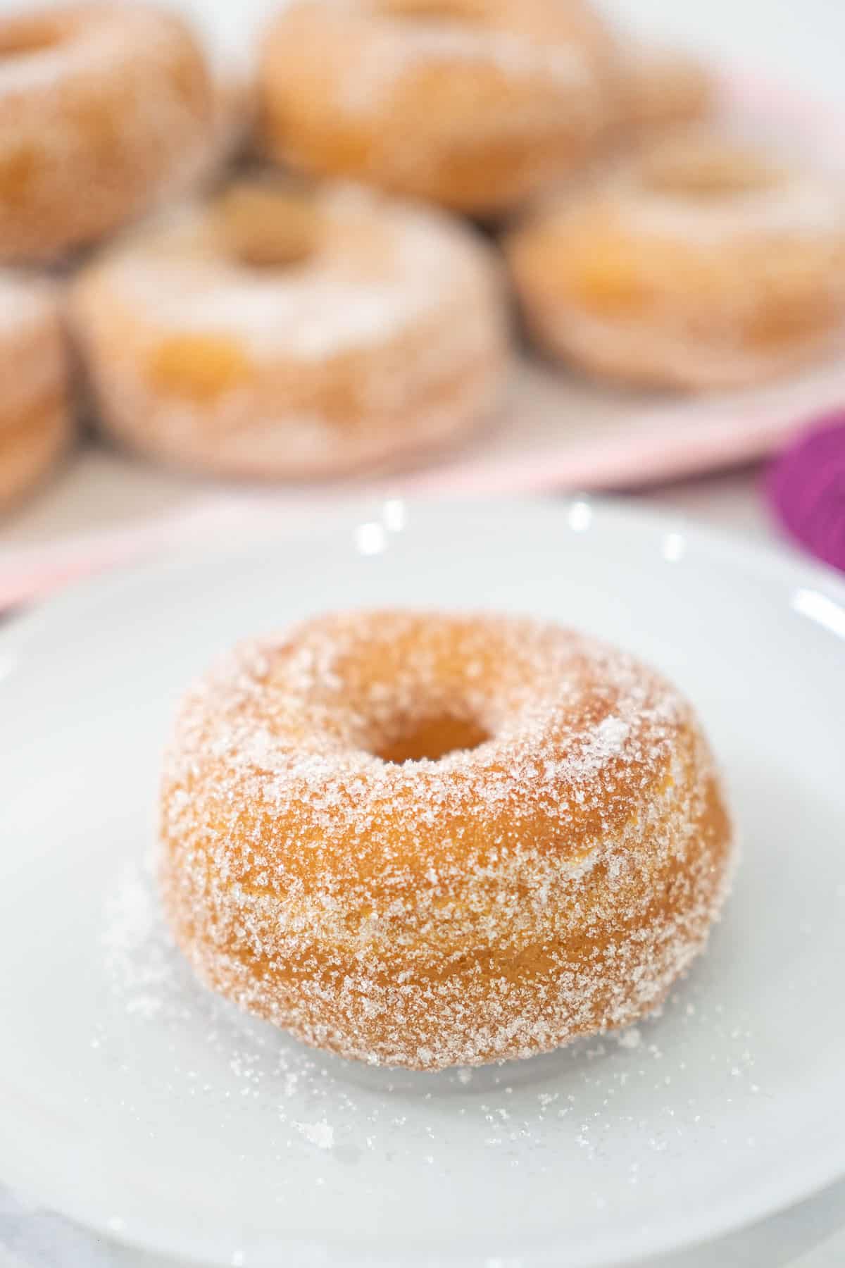 A sugar coated, old fashioned homemade yeast doughnut on a white plate. In the background is a pink tray of doughnuts.