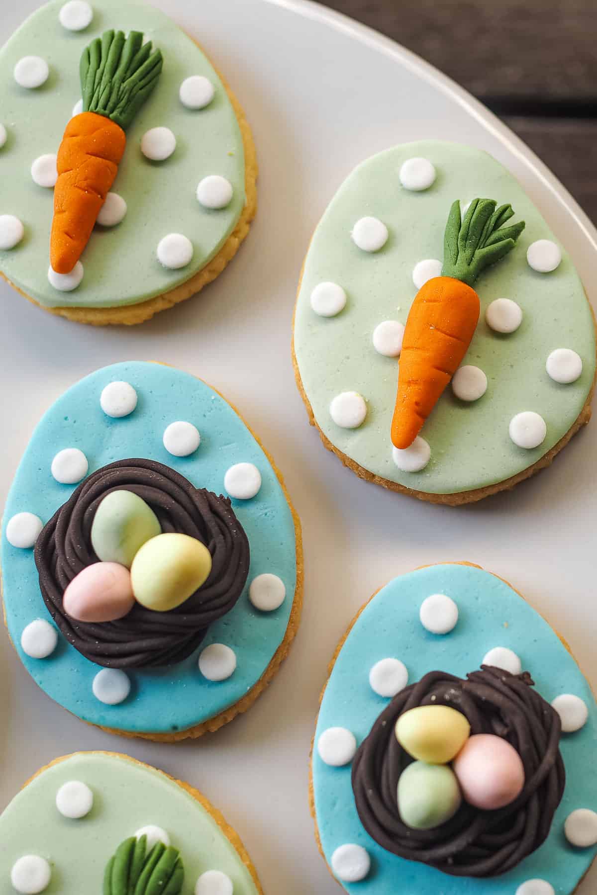 Egg shaped cookies in 2 design. First design with in blue background and white polka dots with 3D bird nest and 3 pastel green, pink and yellow fondant eggs in. The second design is green background with white polka dots and fondant carrot.