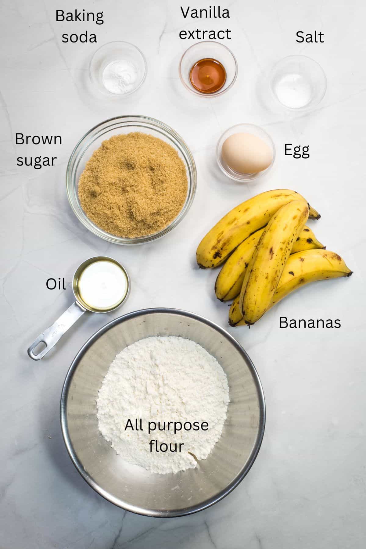 All purpose flour, oil, brown sugar, egg, bananas, baking soda, salt and vanilla extract against a marble background.