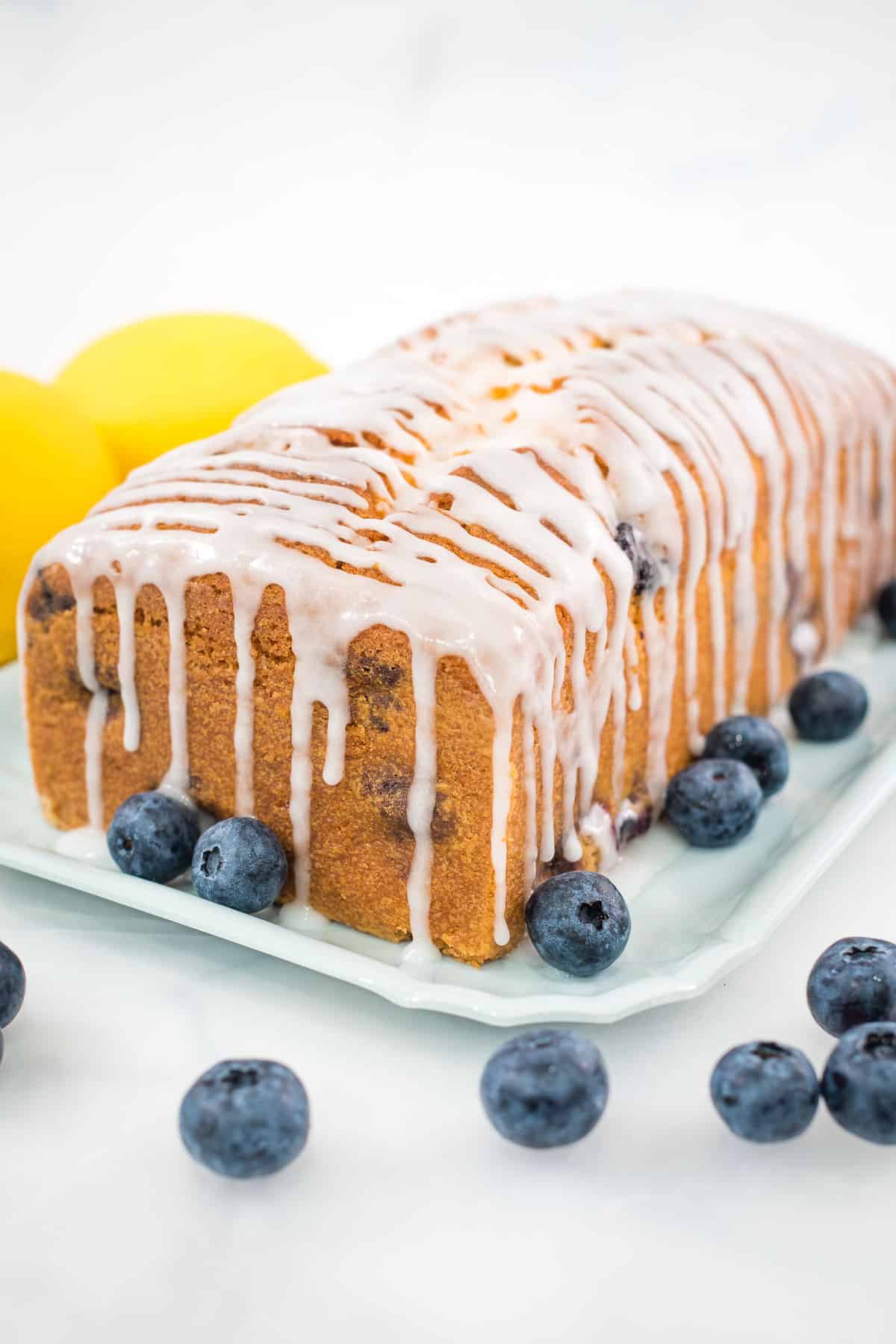 A loaf cake with glaze icing, blueberries and lemons on the side.
