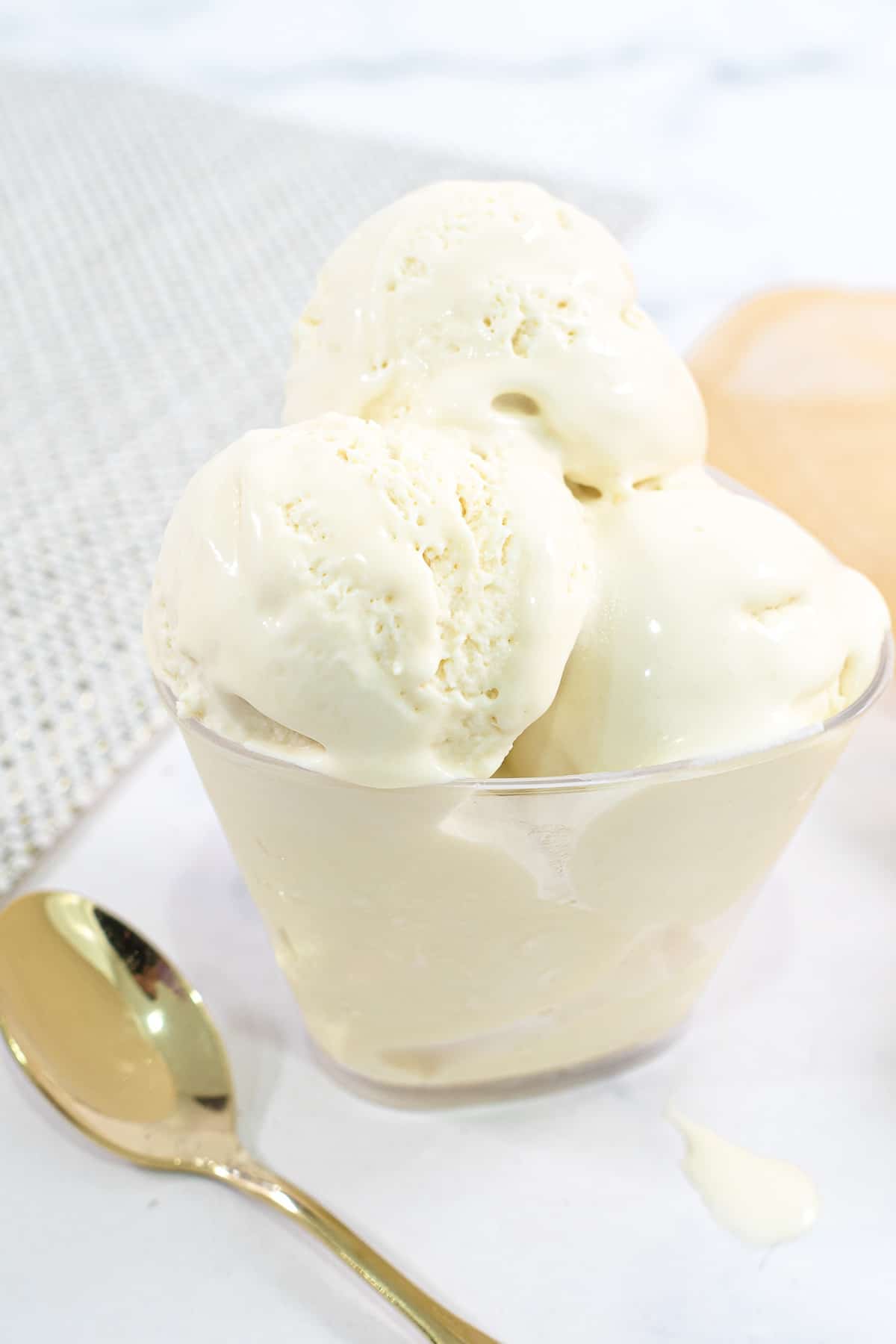 A dessert bowl filled with scoops of vanilla ice cream. A gold spoon is on the side