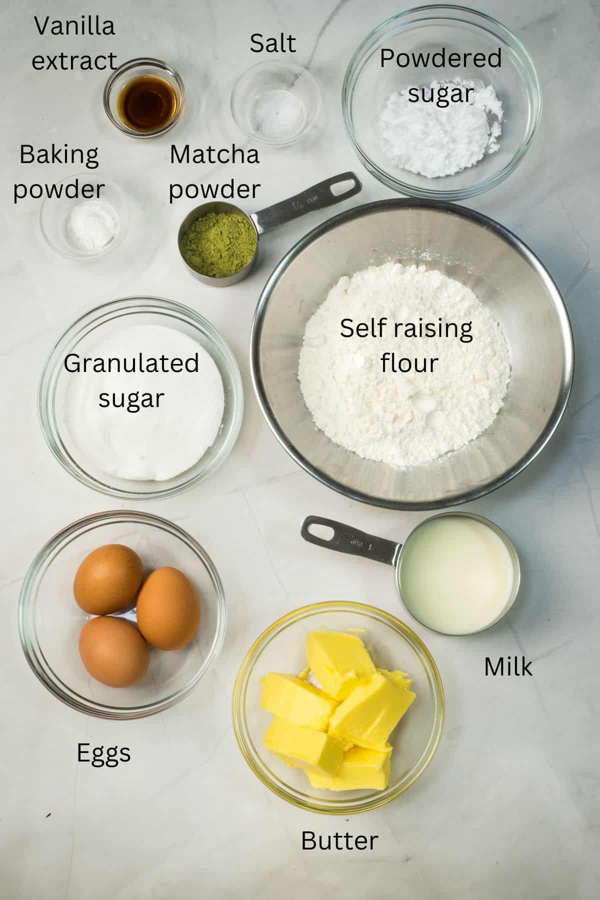 Self raising flour, granulated sugar, eggs, butter, matcha powder, salt, baking powder, milk, vanilla extract and powdered sugar against a marble background.