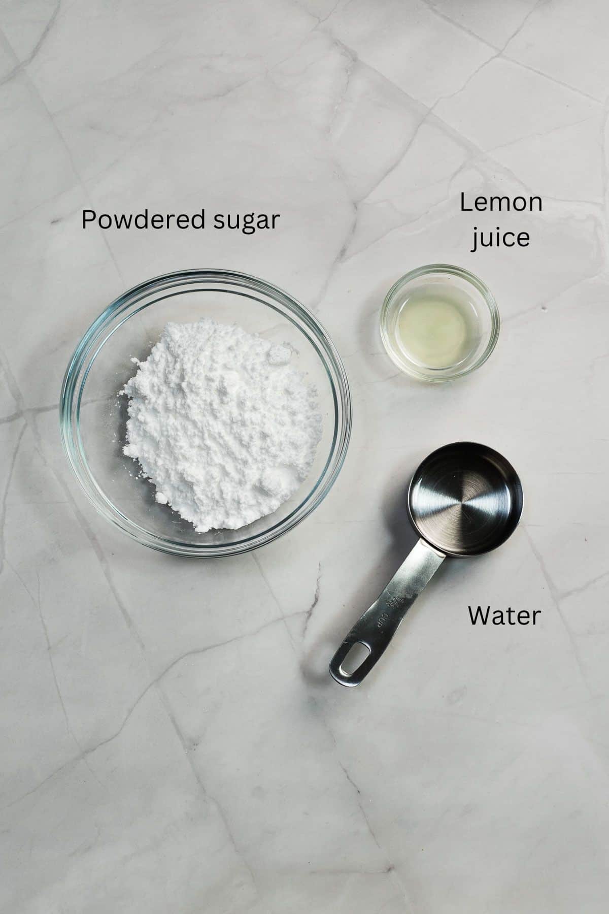 Powdered sugar in a bowl, lemon juice in a bowl and water in a cup against a marble background.