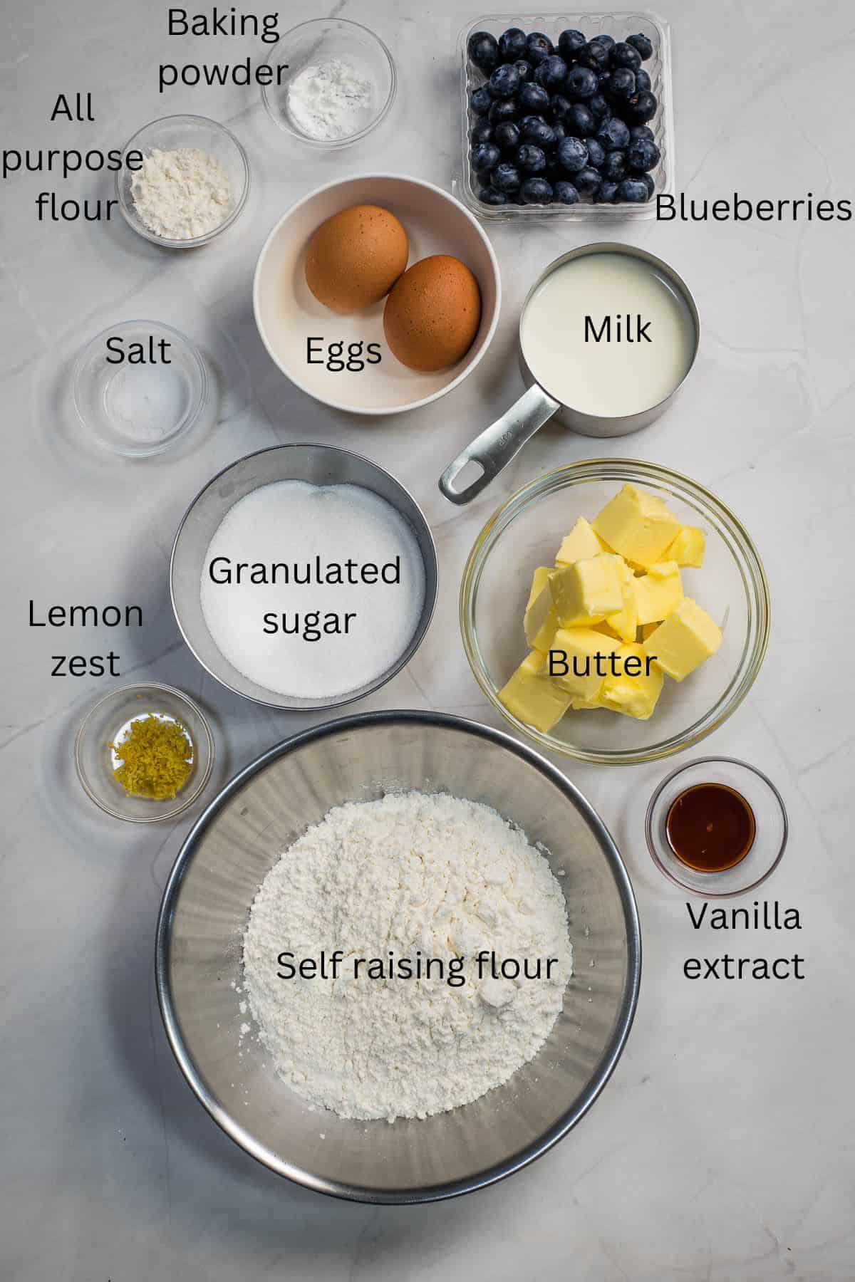 Self raising flour, granulated sugar, butter, eggs, lemon zest, vanilla extract, milk, salt, all purpose flour and blueberries against a marble background.