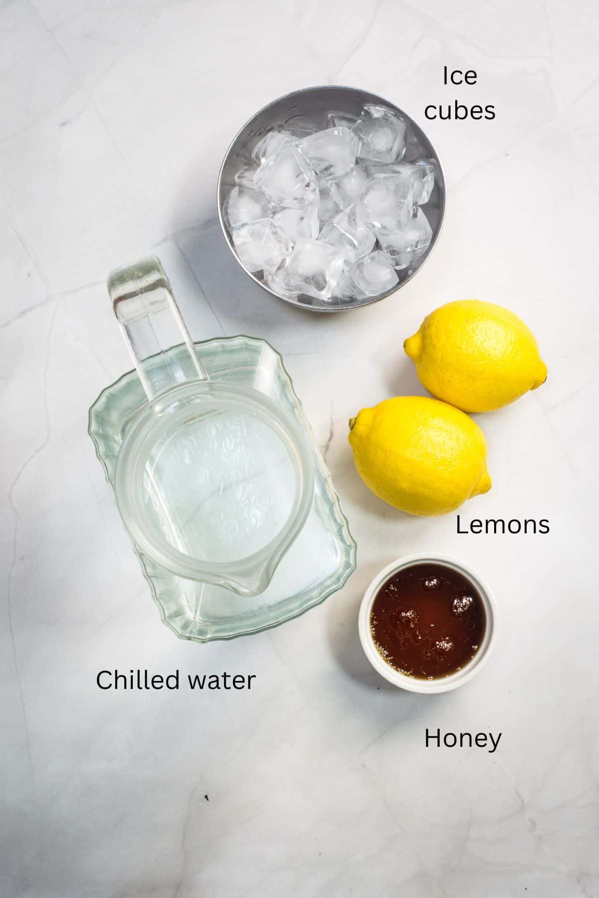 A pitcher of water, 2 lemons, a bowl of honey and a bowl of ice cubes against a marble background.
