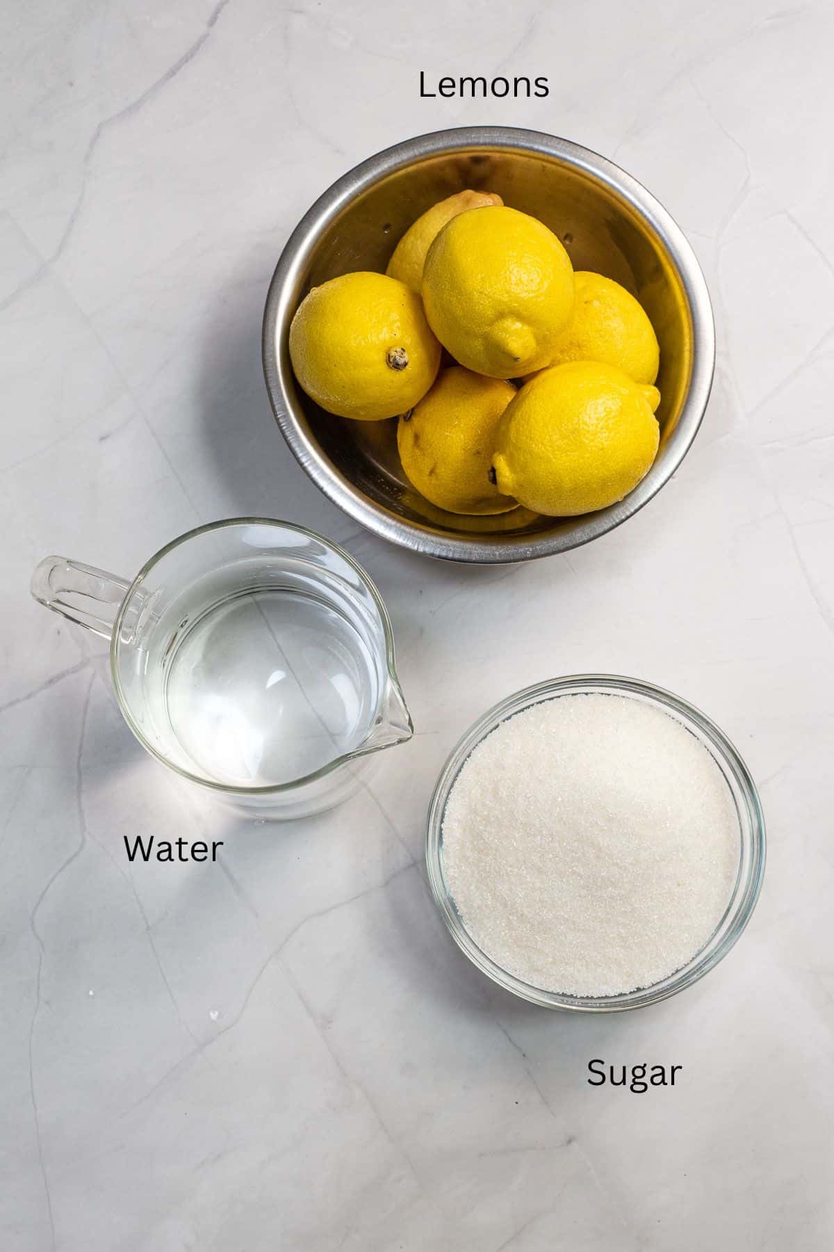 A bowl of lemons, a pitcher of water and a bowl of sugar against a marble background.