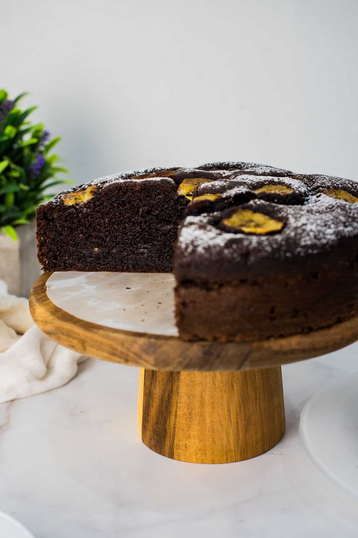 A chocolate cake with banana slices on top, on a wooden cake stand.