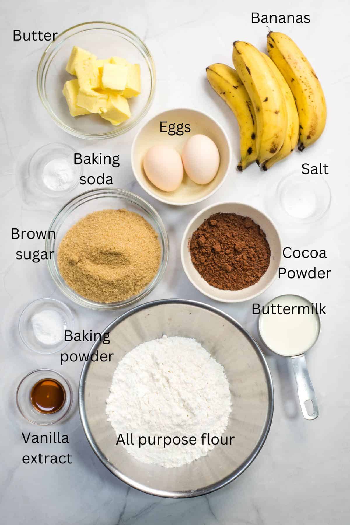 All purpose flour, brown sugar, eggs, cocoa powder, bananas, buttermilk, salt, baking powder, baking soda and vanilla extract in small bowls against a marble background.