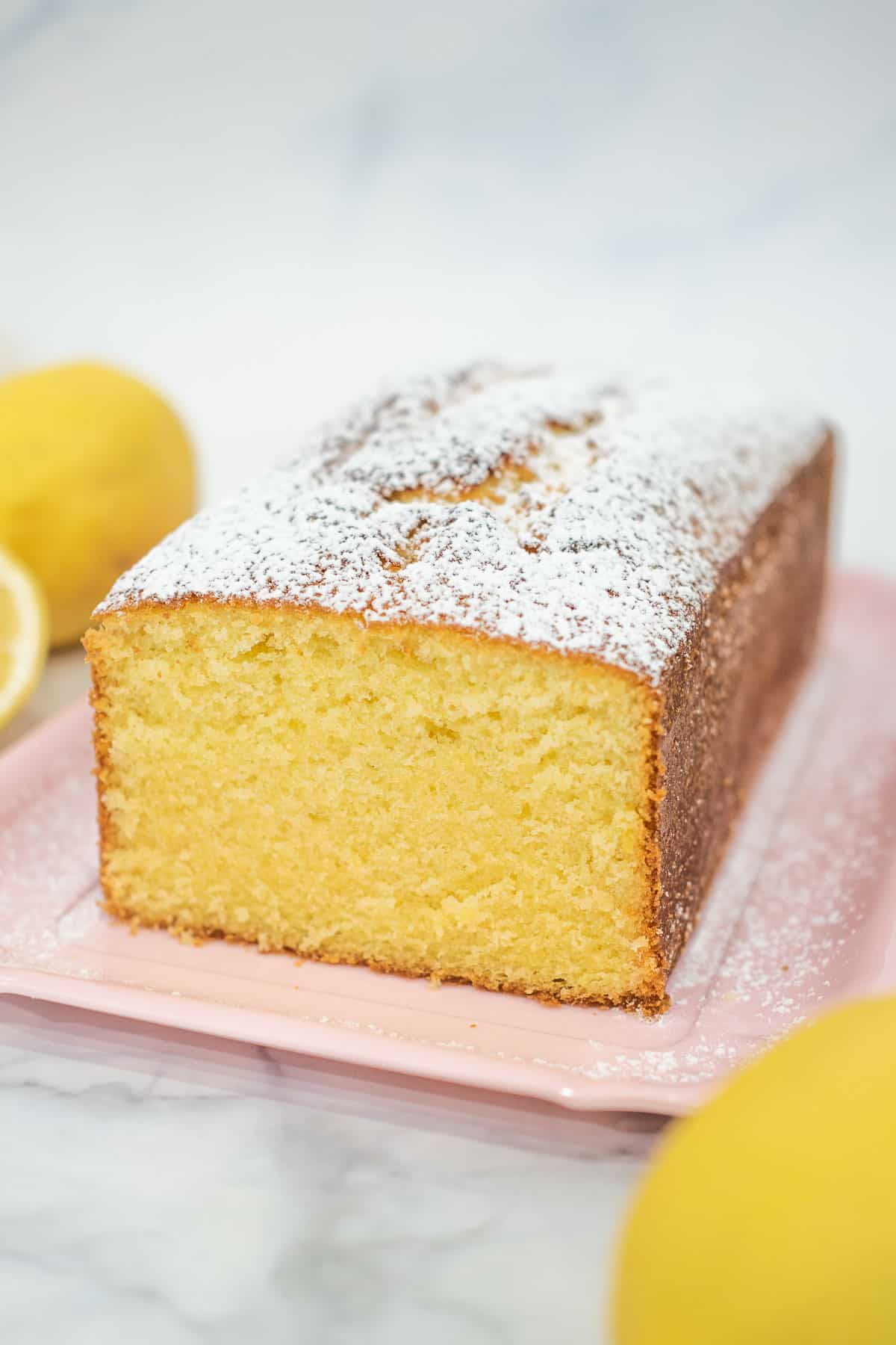 A loaf of lemon cake on a pink tray. Top of the cake is dredged with icing sugar
