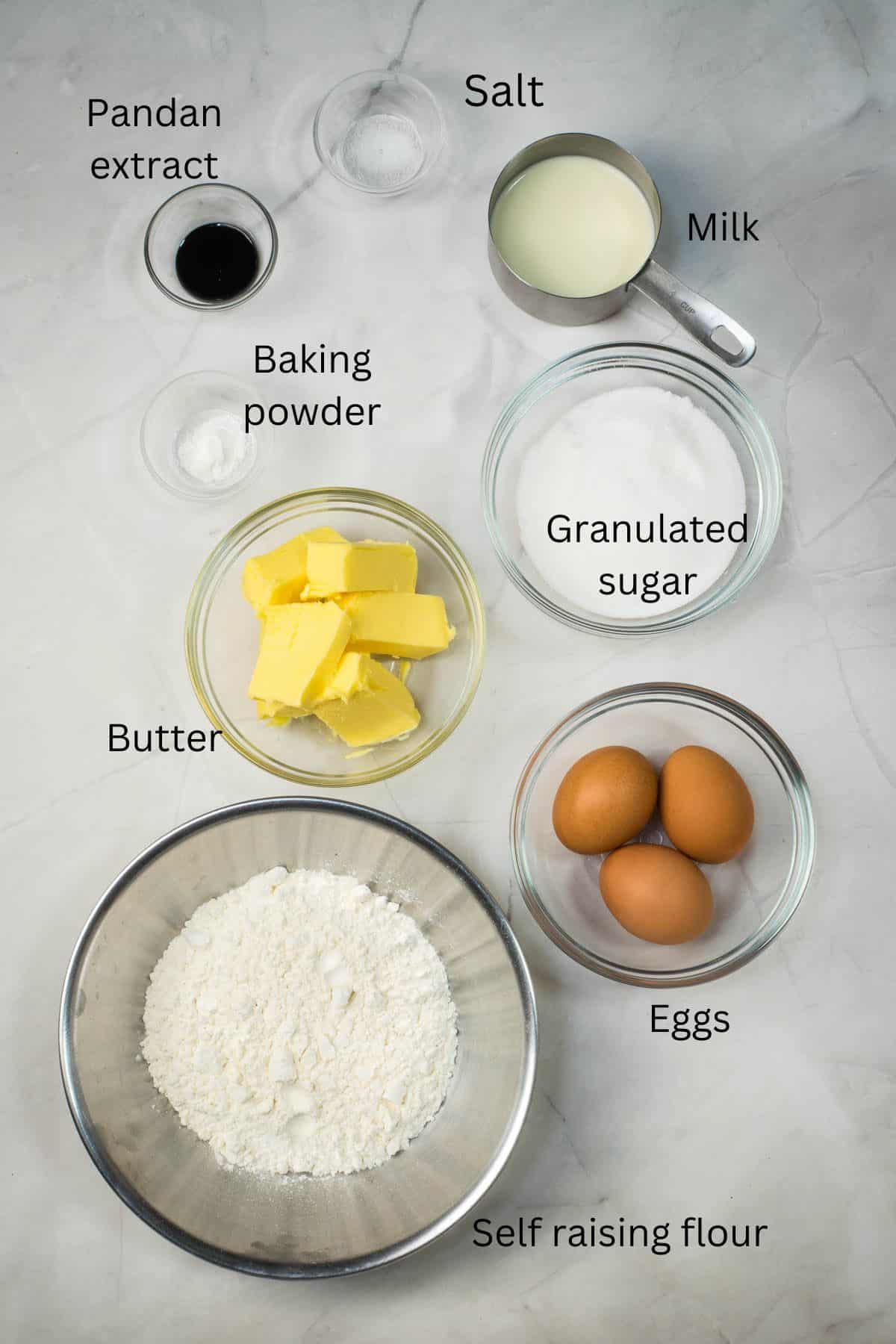 Self raising flour, eggs, butter, granulated sugar, milk, baking powder, salt and pandan extract in bowls against a marble background.