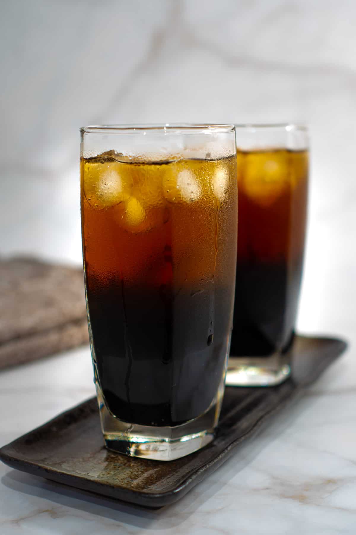 Iced grass jelly tea in 2 tall glasses on a rectangle plate.