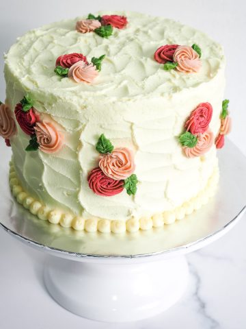 A vanilla cake with pink and peach buttercream rosettes on a white cake stand.