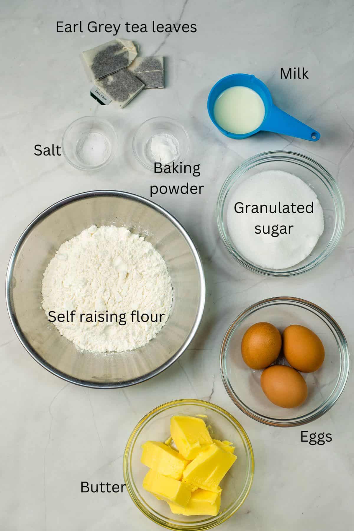 Self raising flour, granulated sugar, butter, eggs, Earl Grey tea bags, salt, baking powder and milk against a marble background.
