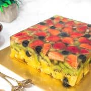 A square cake made of fruits and jelly on a gold cake board.