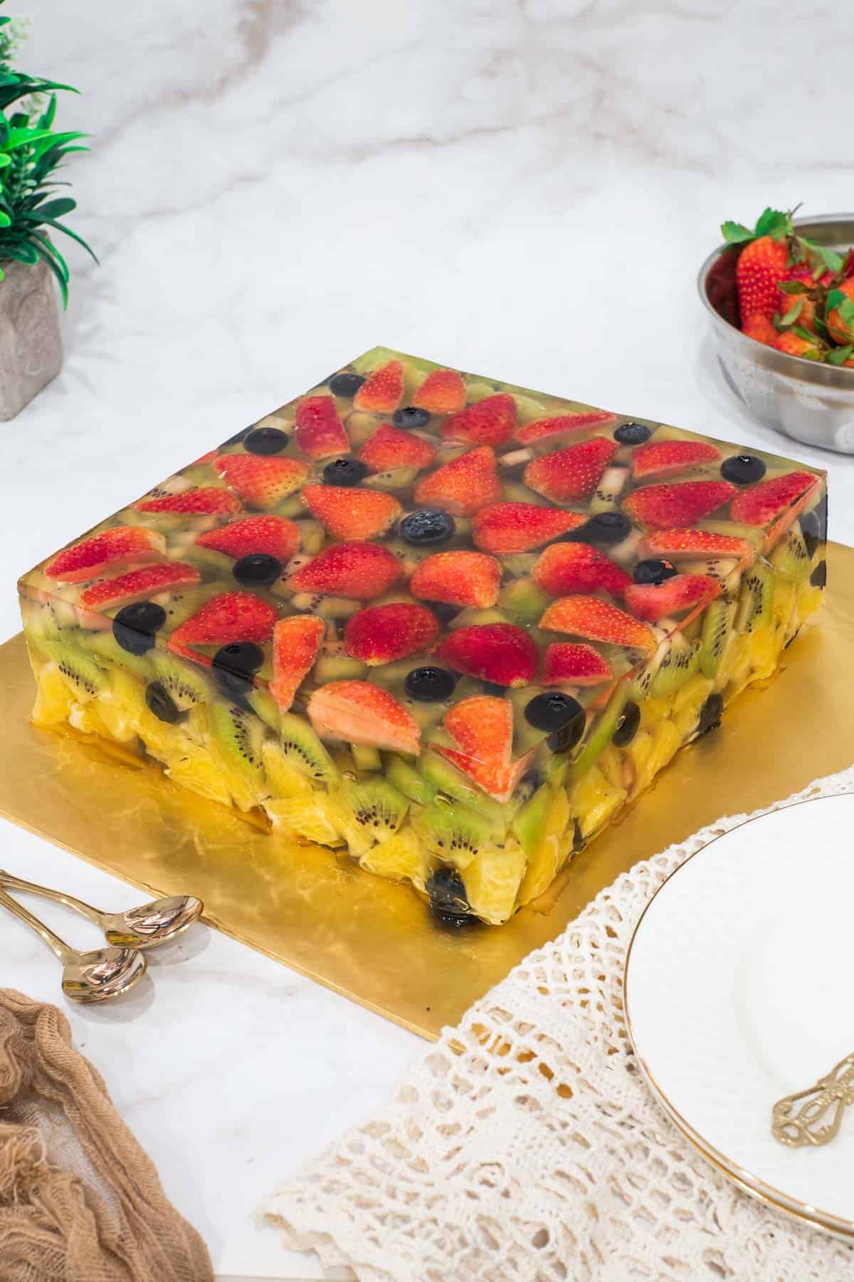 A square cake made of fruits and jelly on a gold cake board.