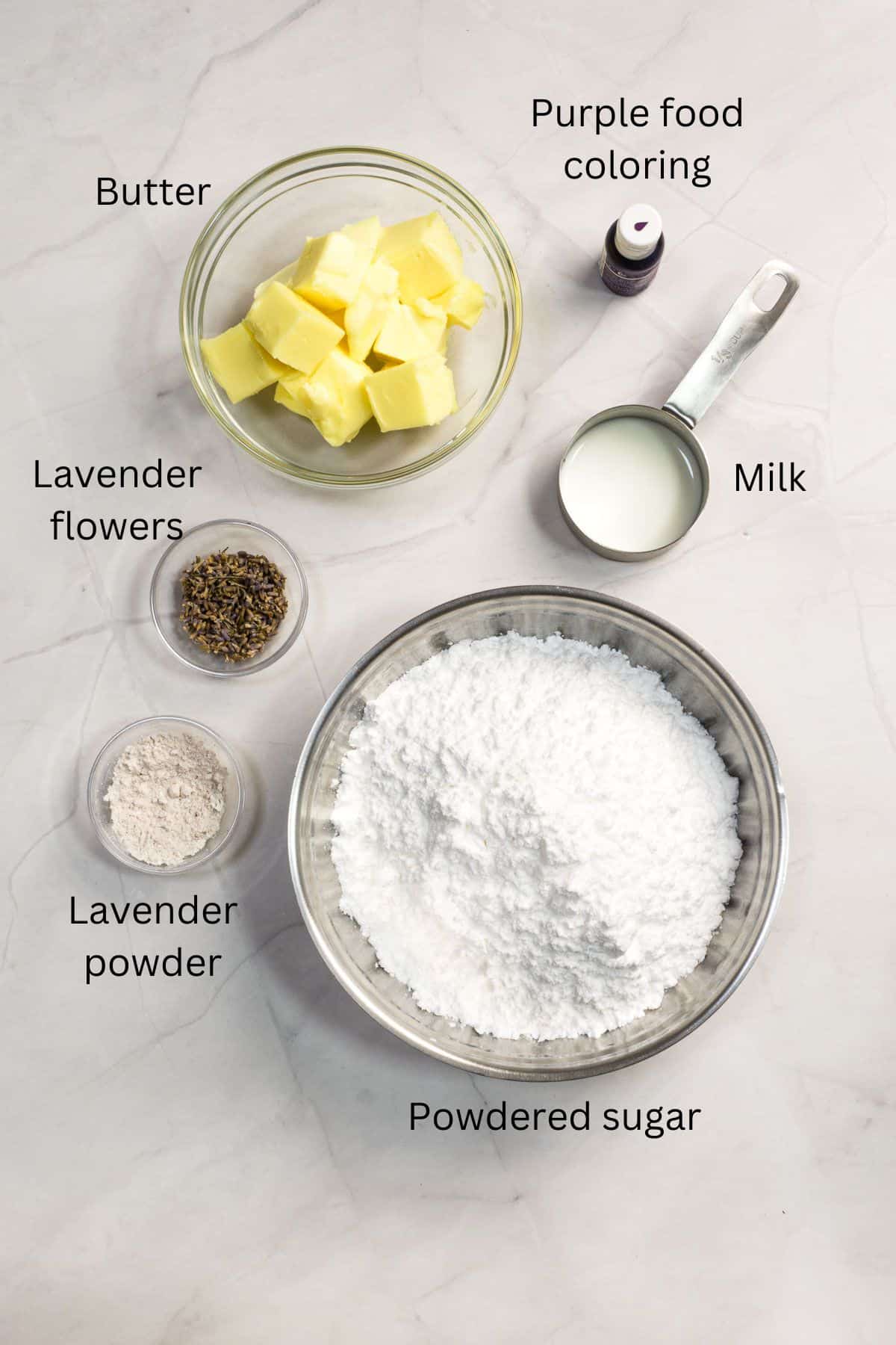 Powdered sugar, butter, lavender buds, lavender powder, purple food coloring and milk in bowls against marble background.