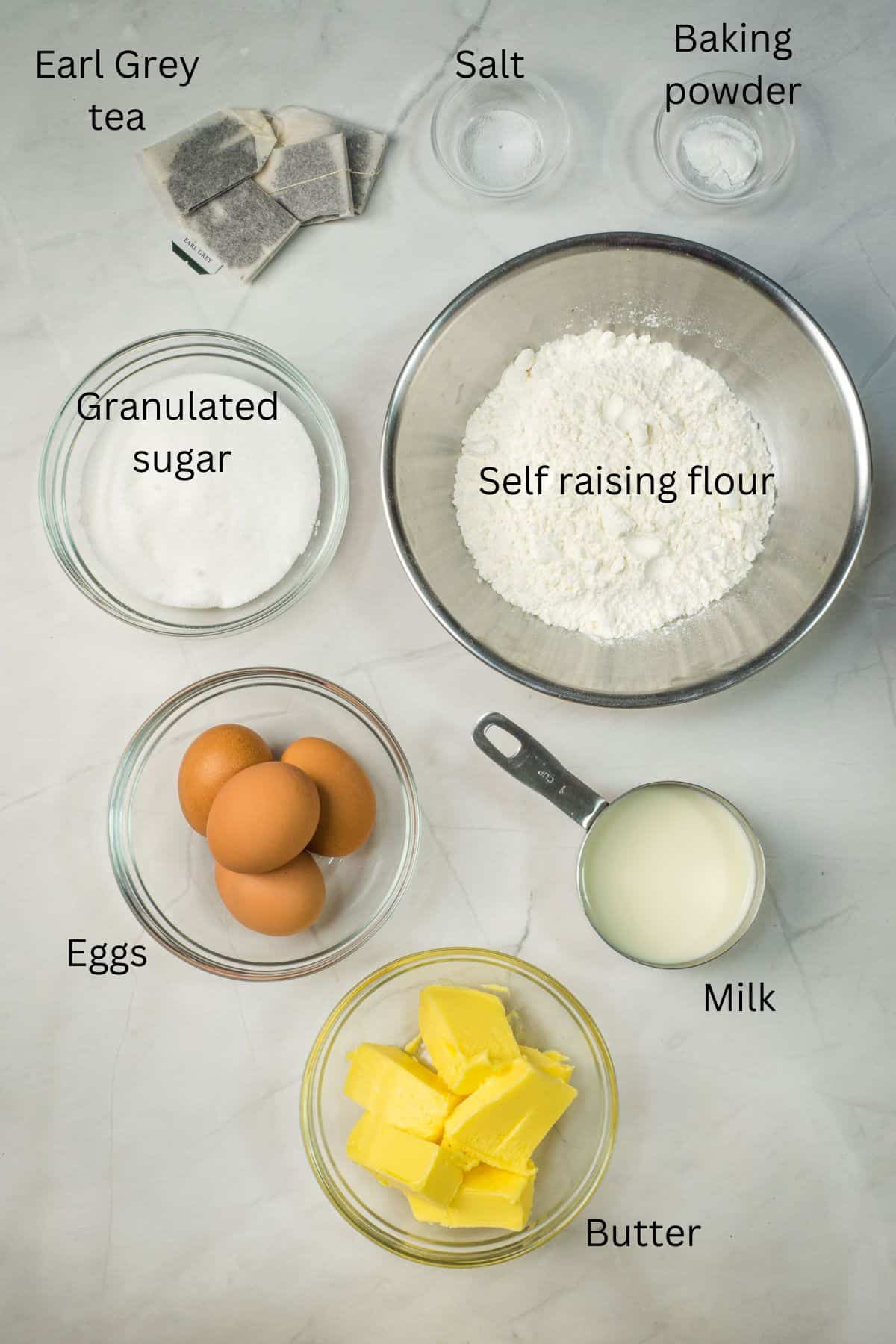 Self raising flour, butter, granulated sugar, eggs, milk, baking powder, salt and earl grey tea in bowls against a marble background.