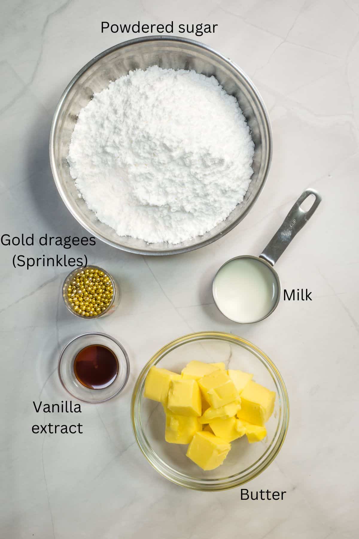 Powdered sugar, butter, vanilla, milk and gold dragees in bowls against a marble background.