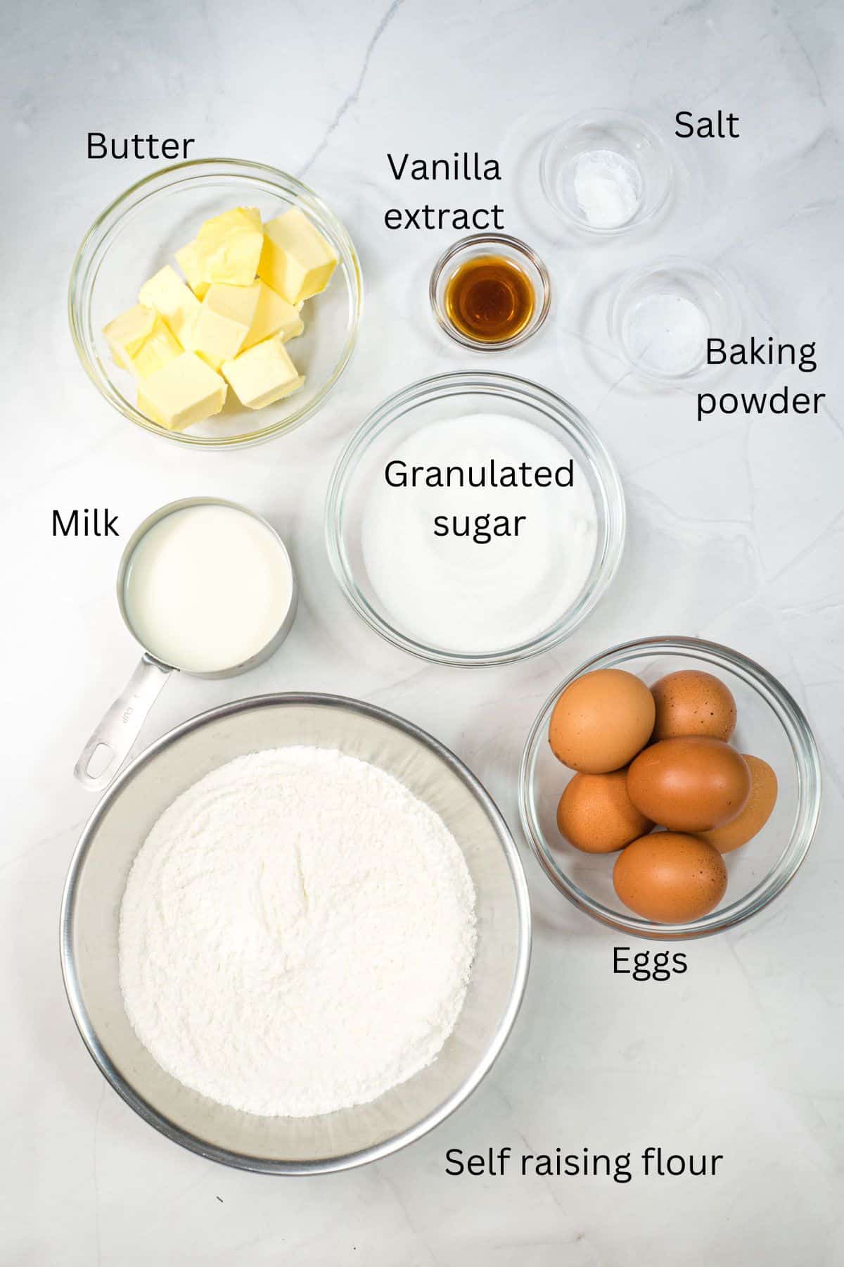 Self raising flour, granulated sugar, baking powder, eggs, milk, vanilla, salt in bowls against a marble background.