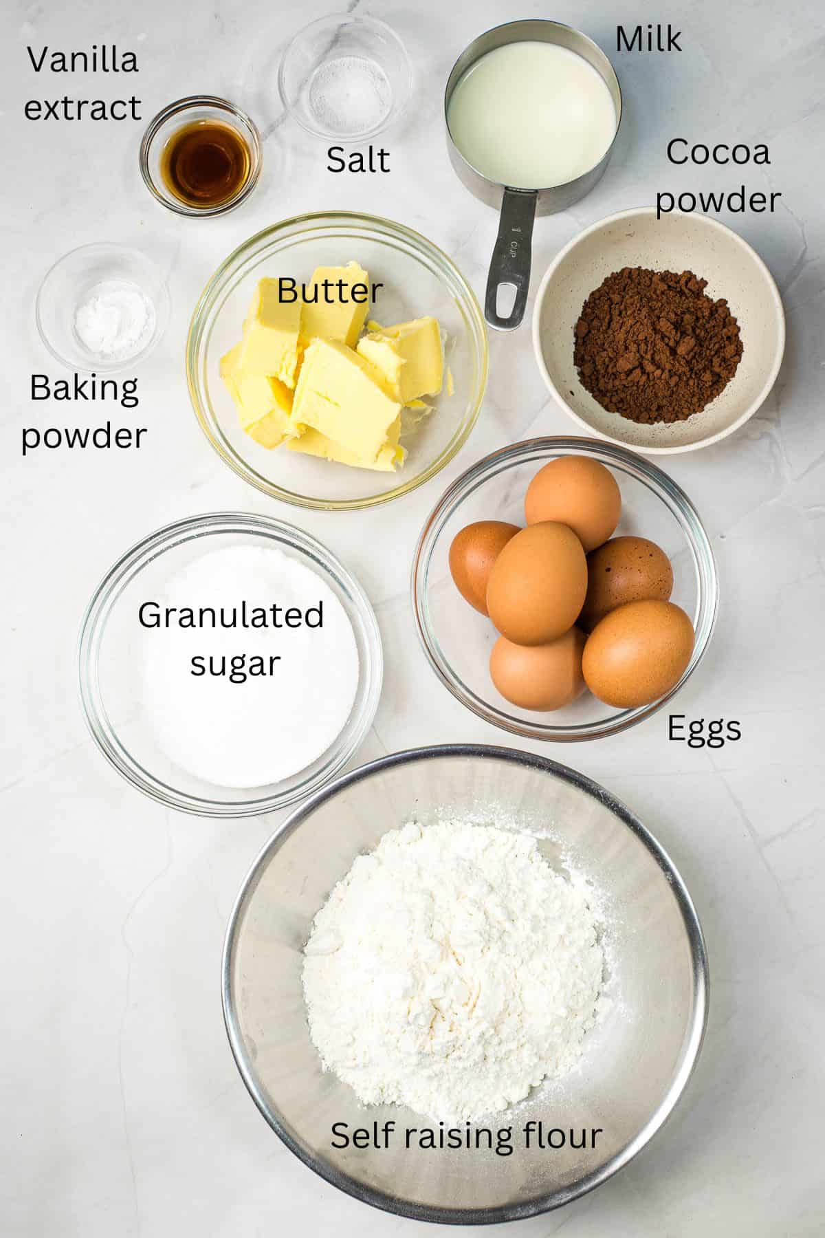 Self raising flour, granulated sugar, eggs, butter, vanilla extract, cocoa powder, salt, baking powder and milk in bowls against marble background.