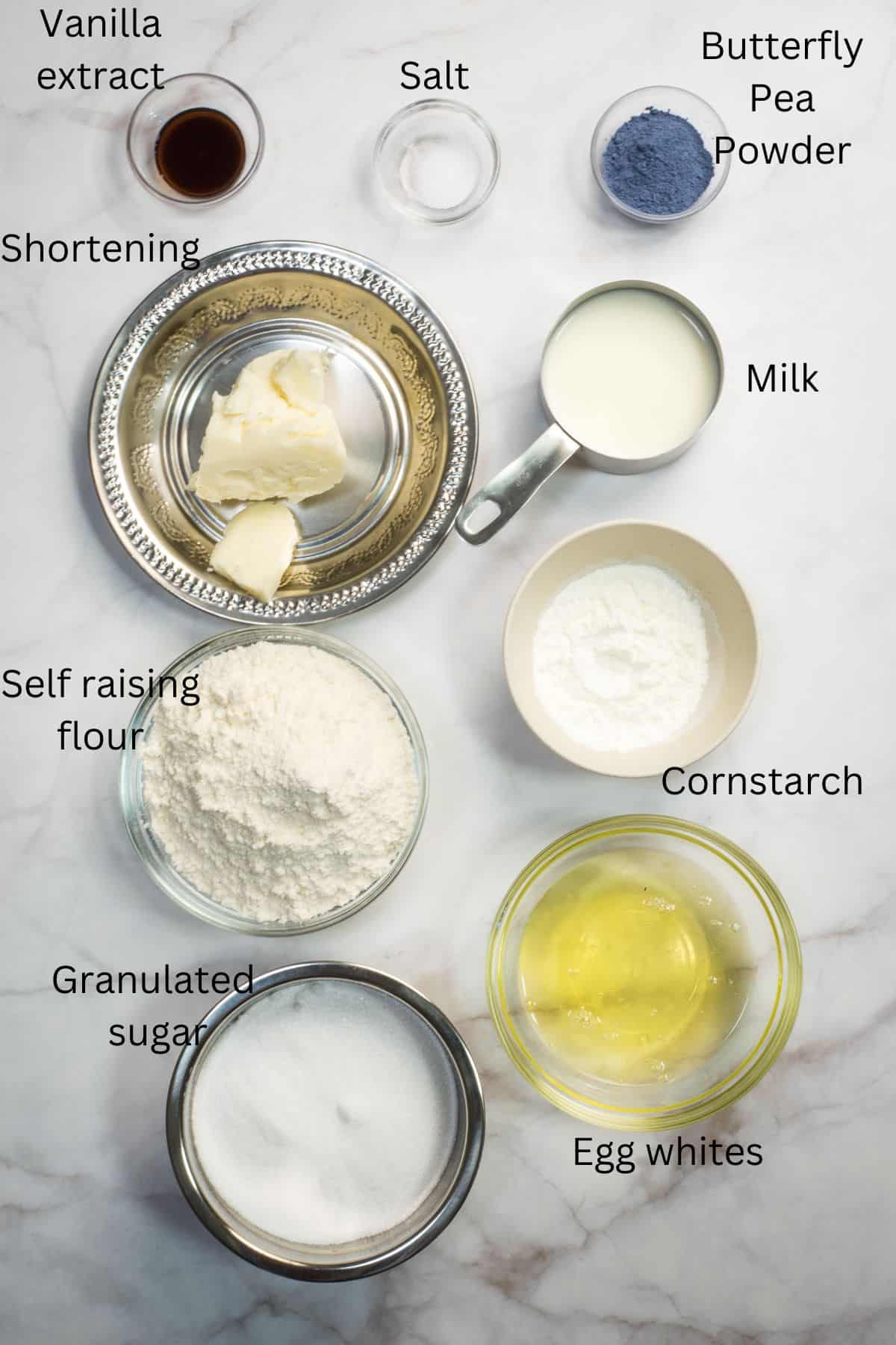 Self raising flour, granulated sugar, egg whites, shortening, milk, cornstarch, butterfly pea flower powder and salt in bowls against a marble background.