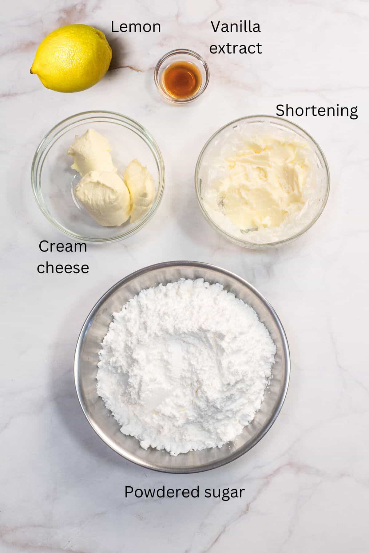 Lemon, vanilla extract, cream cheese, shortening and powdered sugar in bowls against a marble background.