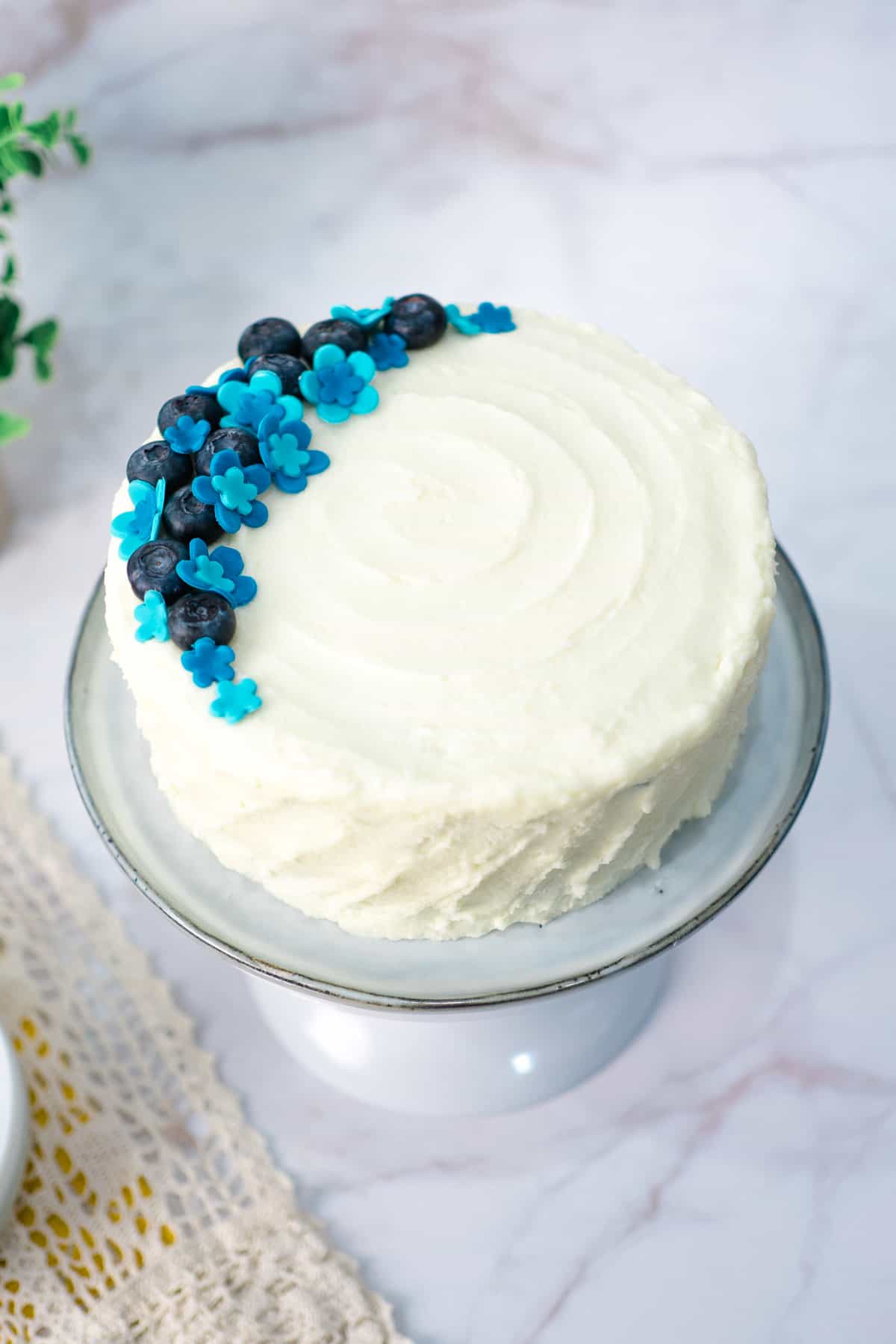 A round cake with white frosting decorated with blue fondant flowers and blueberries.