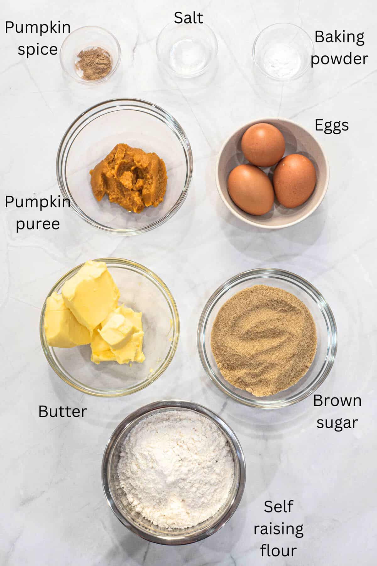 A bowl of flour, brown sugar, butter, pumpkin puree, 3 eggs, baking powder, salt and pumpkin spices against a marble background.