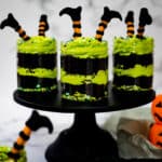 Three jars of chocolate cake layered with green buttercream and decorated with witch legs in orange and black on a black cake stand.