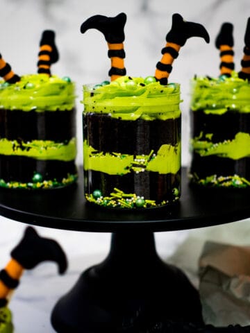 Three jars of chocolate cake layered with green buttercream and decorated with witch legs in orange and black on a black cake stand.