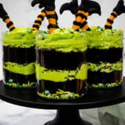 Four jars of chocolate cake layered with green buttercream and decorated with witch legs in orange and black on a black cake stand.