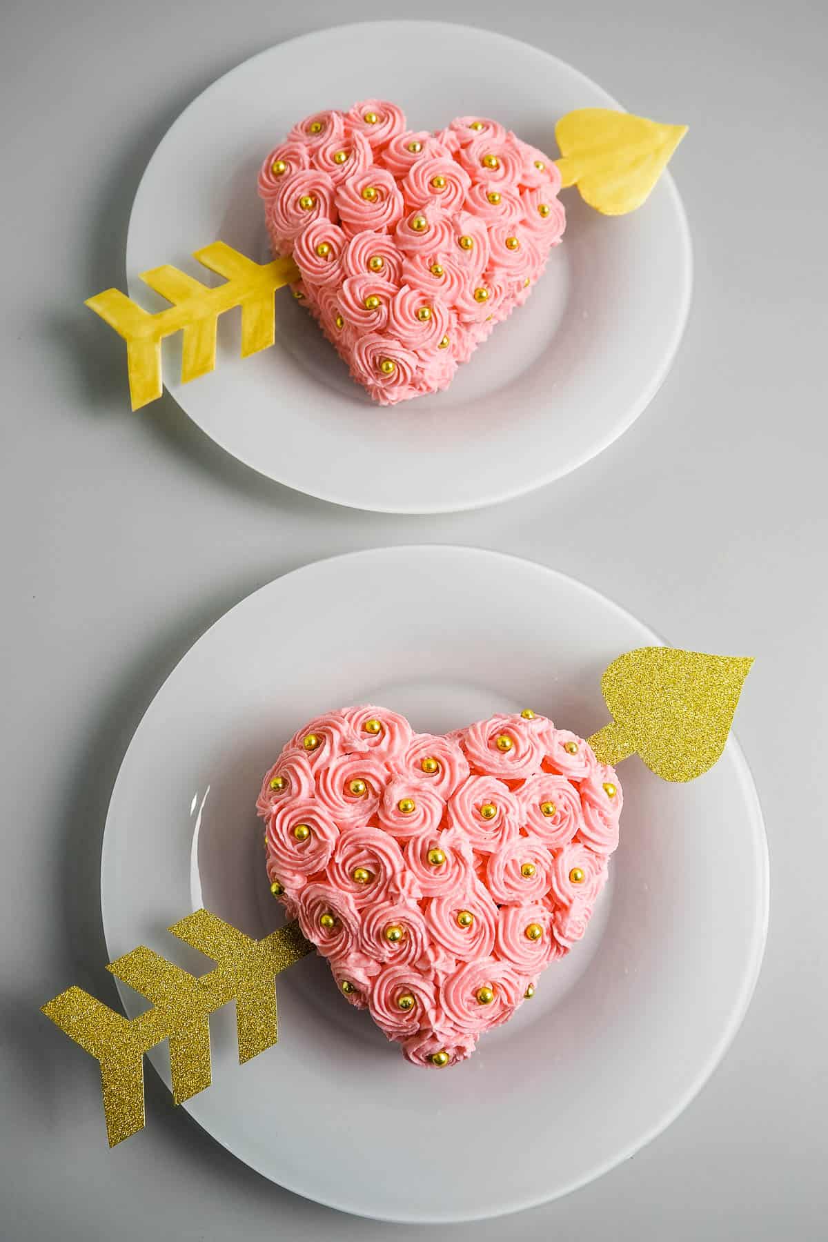 Two heart shaped cakes with pink buttercream rosettes frosting and gold arrows on white plates.