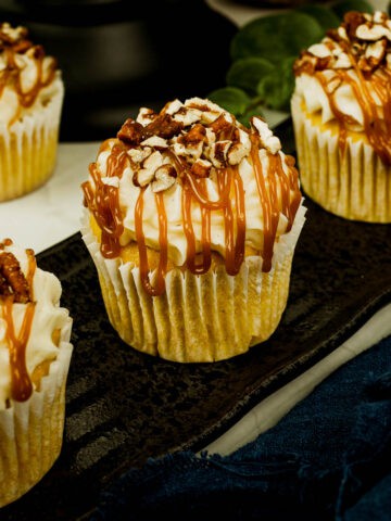 Pumpkin cupcakes with white frosting, caramel drizzle and chopped pecans on a brown plate.