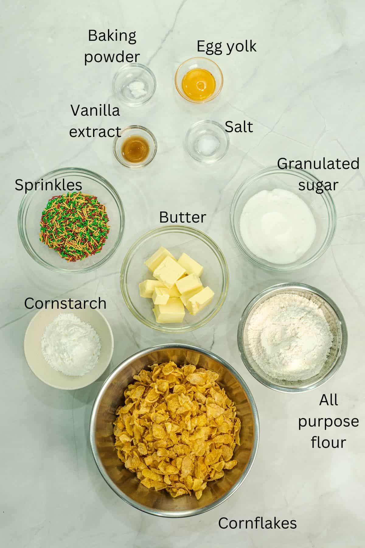 Cornflakes, cornstarch, flour, butter, sugar, sprinkles, vanilla, salt, baking powder and egg yolk in bowls against a marble background.