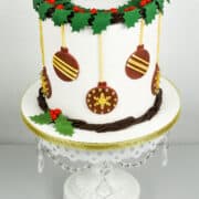 A round cake with holly leaves and baubles decoration on a white cake stand.
