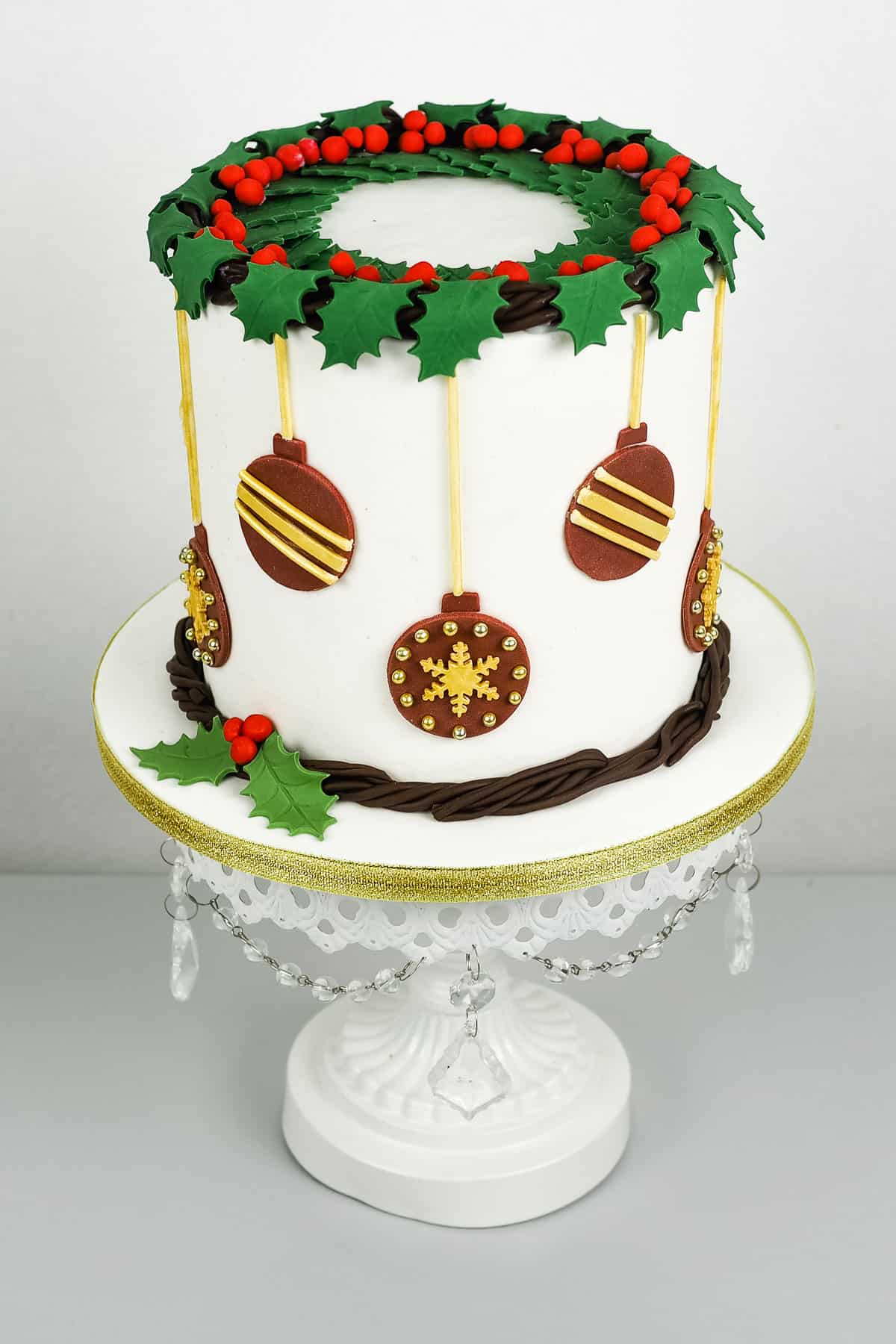 A round cake with holly leaves and baubles decoration on a white cake stand.