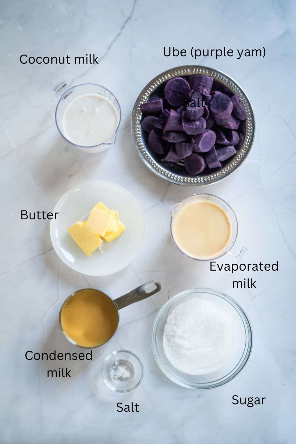 Plates and bowls of ube, coconut milk, butter, evaporated milk, condensed milk, sugar and salt against a marble background.