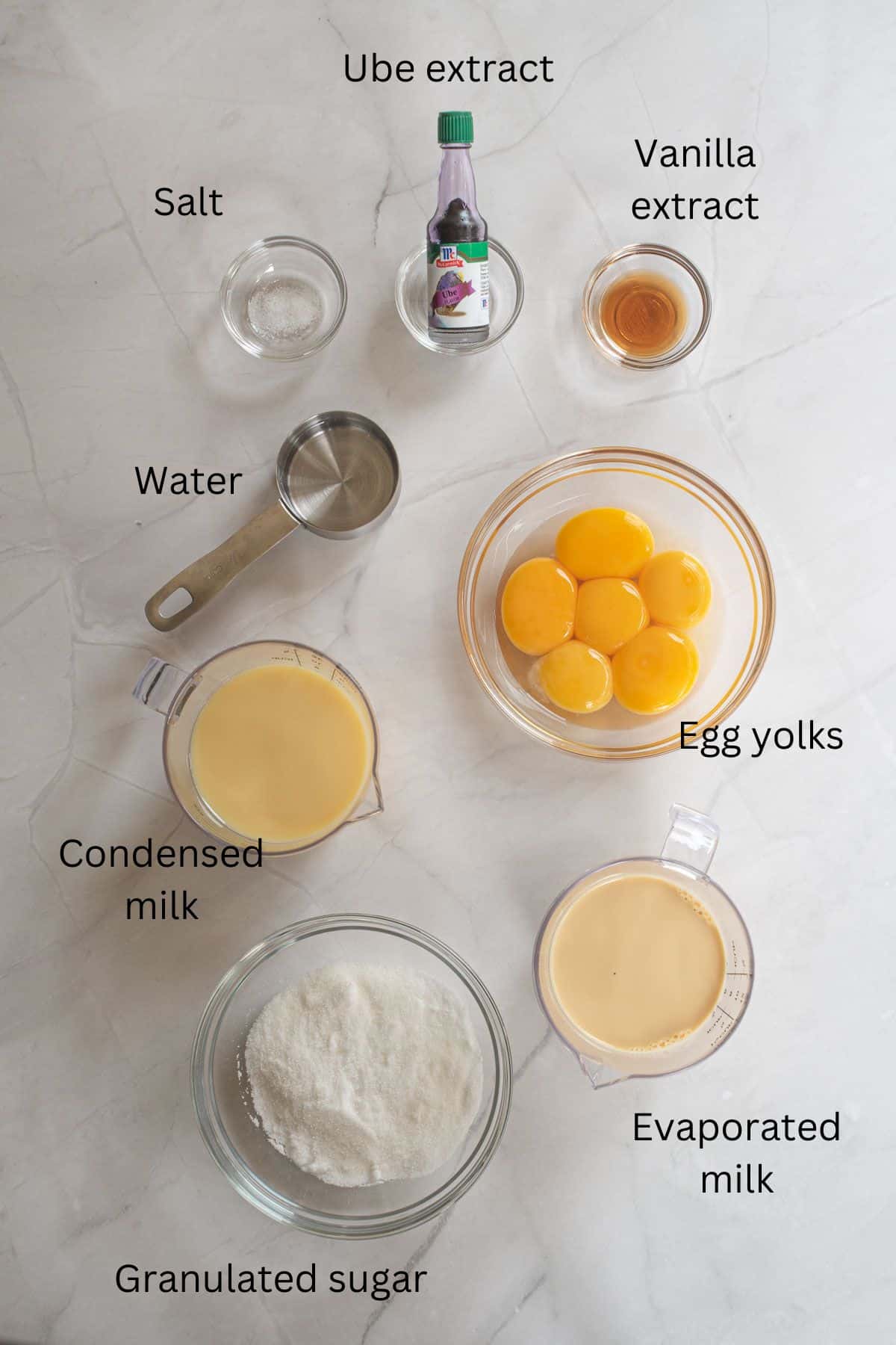 Egg yolks, condensed milk, evaporated milk, vanilla extract, ube extract, sugar, salt and water against a marble background.