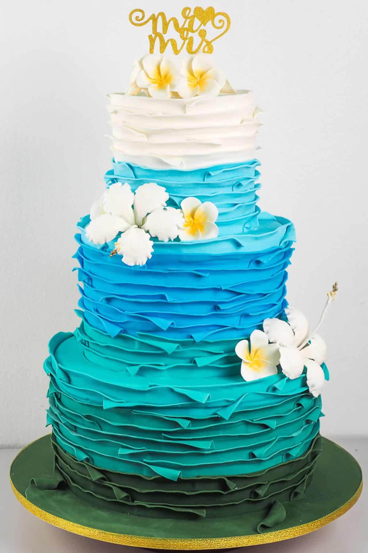 A 3 tier cake decorated in blue and turquoise and plumeria and hibiscus, with and Mr and Mrs cake topper.